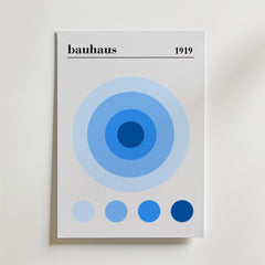 Bauhaus Blue Circles Poster by Bozetto har "bauhaus" och "1919" högst upp, koncentriska blå cirklar och en rad med fem blå cirklar på en ren vit bakgrund i en minimalistisk konstaffisch stil. Tryckt på premiumpapper.
