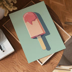 The no-9 green poster, featuring a vibrant popsicle with pink topping, rests on a wooden table beside a book and tray in a warmly lit room with herringbone wood flooring.