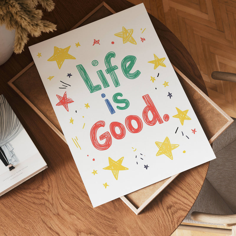 The no-9 poster, featuring large playful "Life is Good" text surrounded by hand-drawn stars and doodles, sits on a brown tray atop a wooden table near a book and chair.