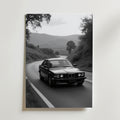 Vintage BMW Poster by Bozetto: Svartvitt foto av en klassisk sedan som kör på en slingrande väg genom kullar, omgiven av träd och berg, tryckt på premiumpapper med en lyxig matt finish.