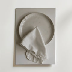 En Bozetto White linen dinner napkin Poster visar en vit keramiktallrik som toppas med en fransig vit linneservett, allt mot en vit bakgrund med en lyxig matt finish.